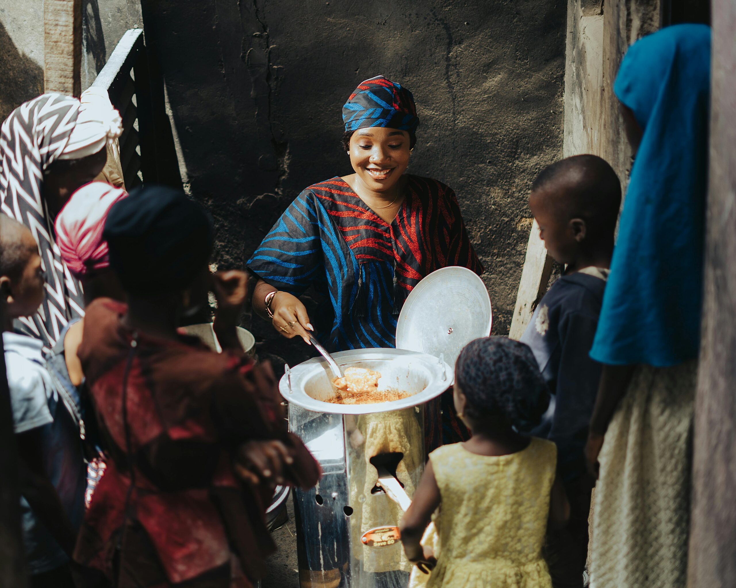 A woman serves food from a large pot to a group of children outdoors in an African community.